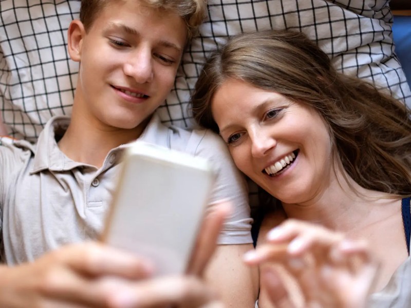 Mutter liegt neben Teenagersohn im Bett und beide schauen sich etwas Lustiges auf dem Smartphones des Sohnes an.