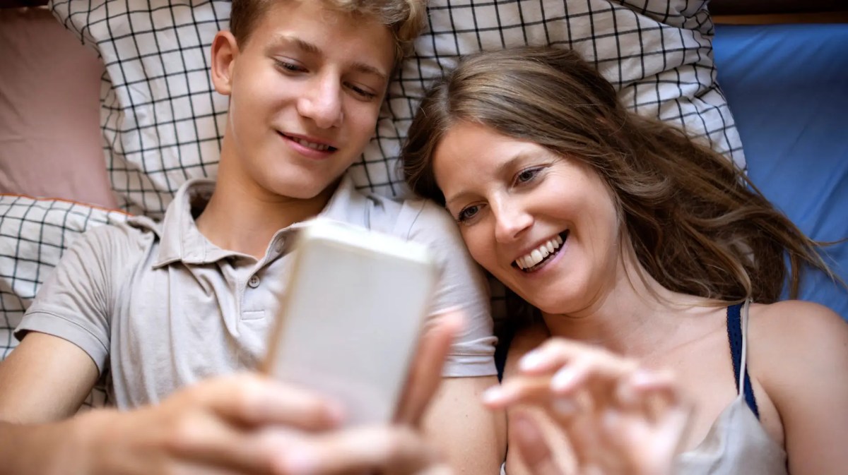Mutter liegt neben Teenagersohn im Bett und beide schauen sich etwas Lustiges auf dem Smartphones des Sohnes an.