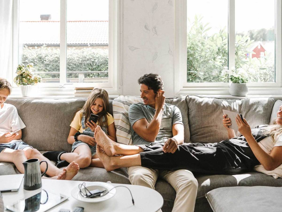 Vierk&ouml;pfige Familie sitzt auf dem Sofa, alle haben ein digitales Ger&auml;t in den H&auml;nden.