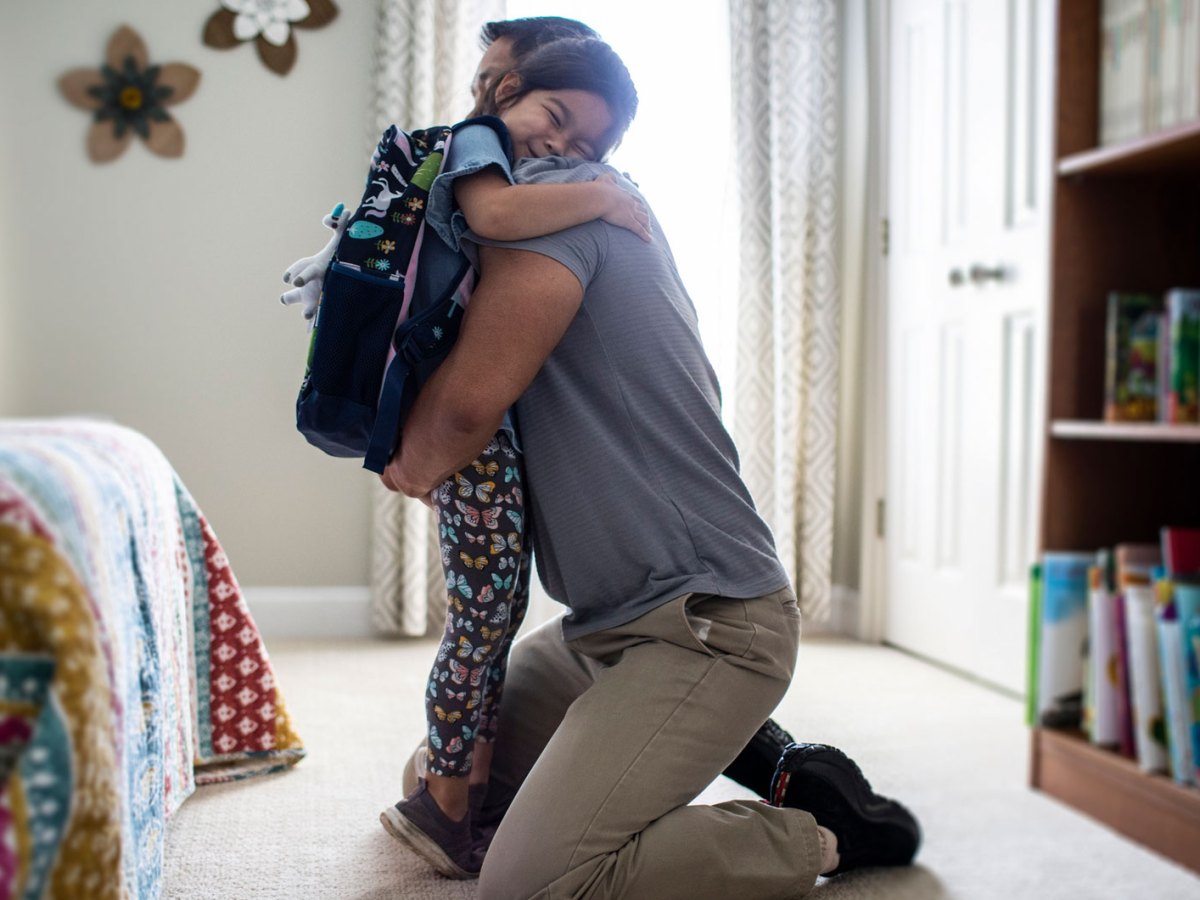 Vater umarmt seine Tochter, die einen Rucksack trägt, im Schlafzimmer.