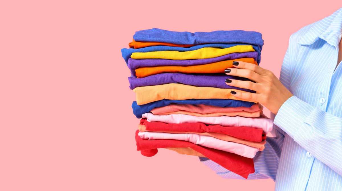 Junge Frau mit einem Stapel bunter Kleidung auf rosa Hintergrund, Nahaufnahme.