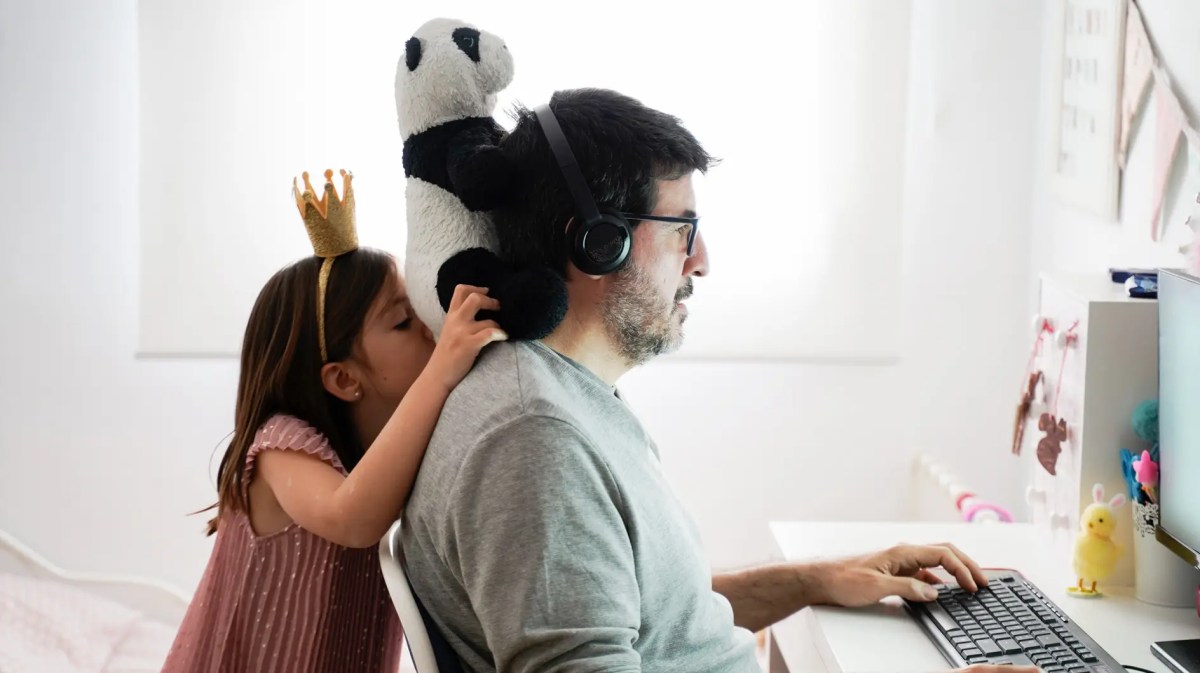Vater am Computer, seine Tochter dahinter mit einem Plüschtier.