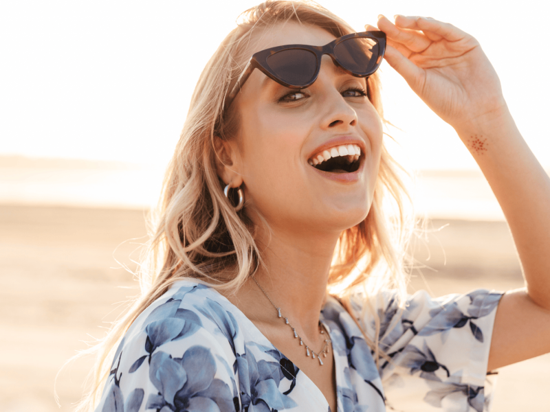 Frau mit Sonnenbrille an Strand lächelt in Kamera.