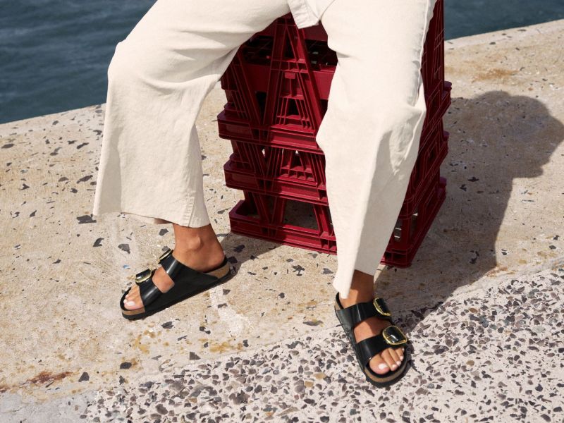 Frau in weißer Jeans und schwarzen Birkenstock-Sandalen sitzt auf roten Kisten am Meer