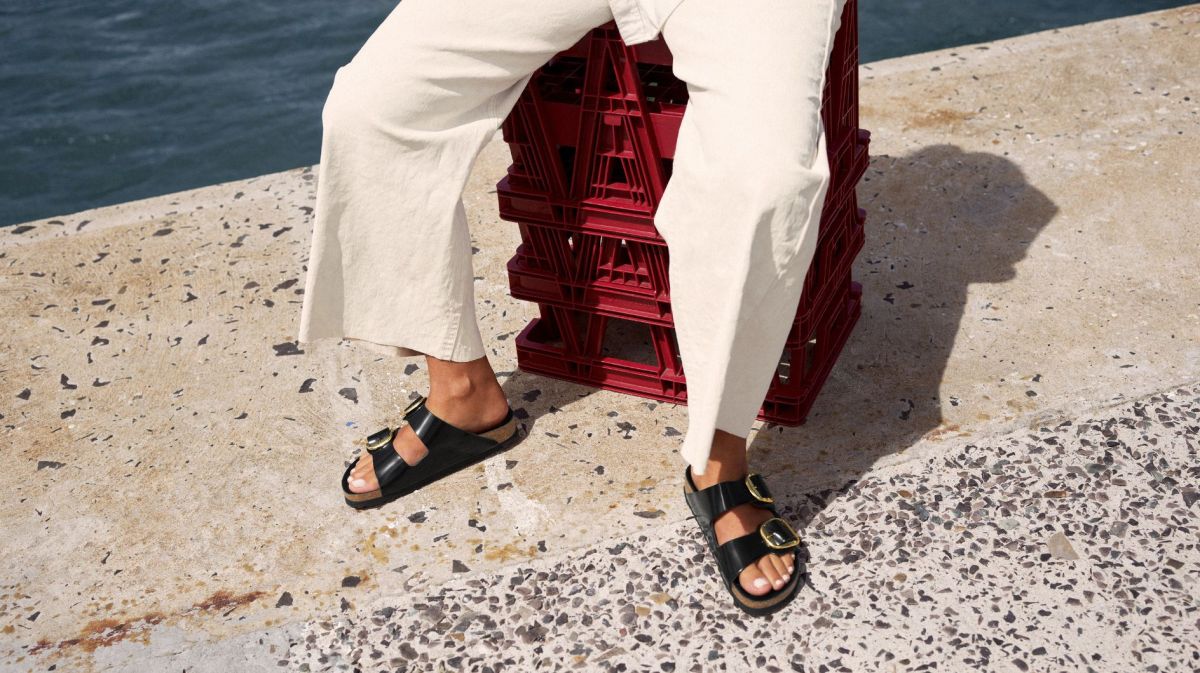 Frau in weißer Jeans und schwarzen Birkenstock-Sandalen sitzt auf roten Kisten am Meer