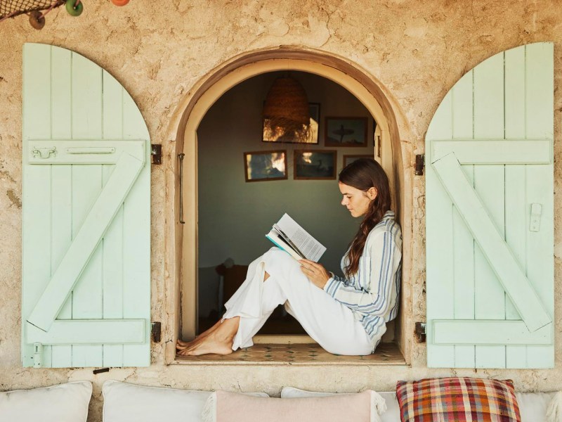 Junge Frau sitzt am Fenster und liest ein Buch, Urlaubsatmosphäre