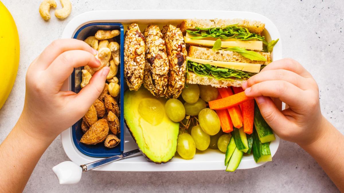 Kind sitzt vor einer Brotdose gefüllt mit Brot, Gemüse und Obst und isst davon.