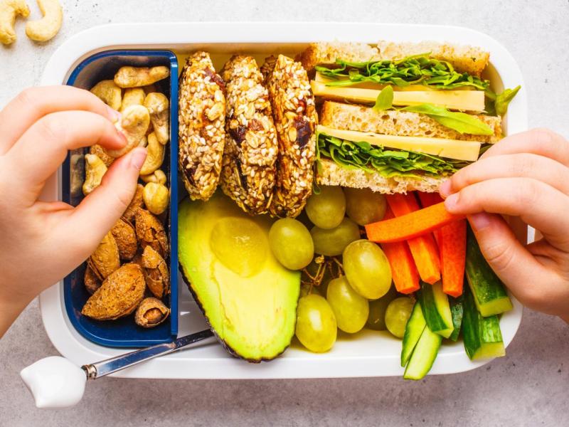 Kind sitzt vor einer Brotdose gefüllt mit Brot, Gemüse und Obst und isst davon.