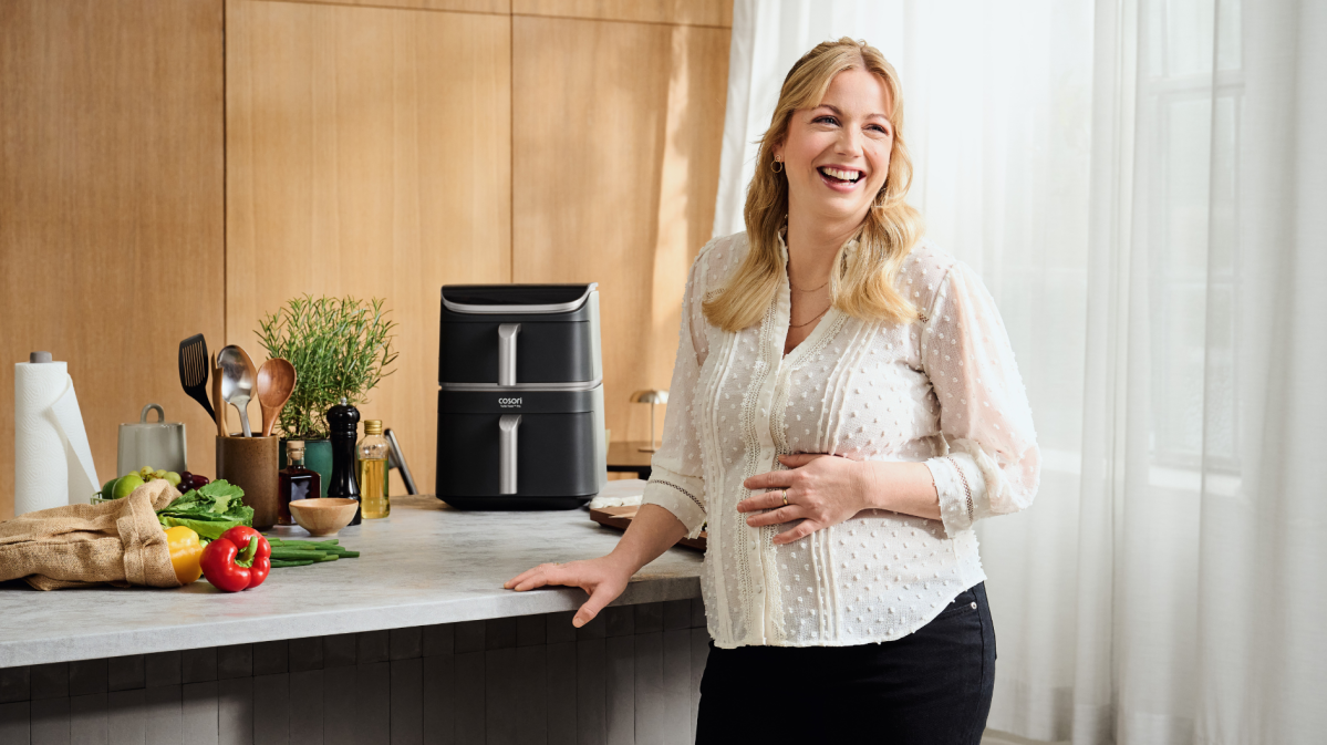 Junge blonde Frau steht am Tisch, neben ihr eine Heißluftfritteuse