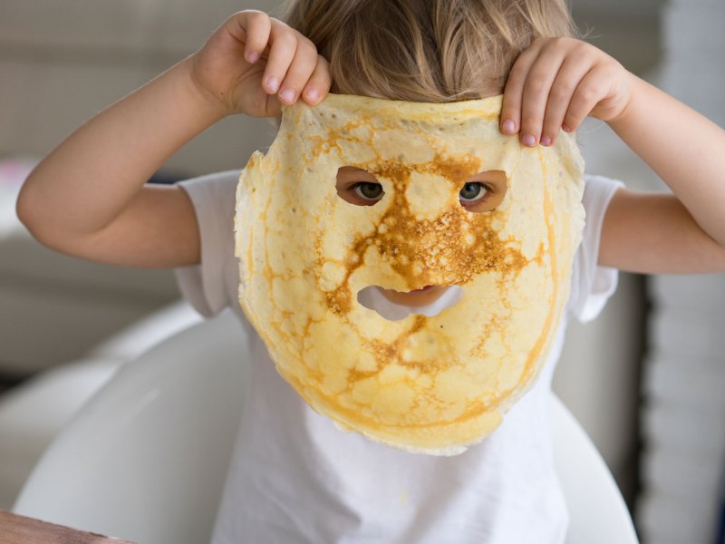 Nahaufnahme eines kleinen Kindes, das am K&uuml;chentisch sitzt und sich einen Pfannkuchen vors Gesicht h&auml;lt, in dem L&ouml;cher f&uuml;r Mund und Augen sind.