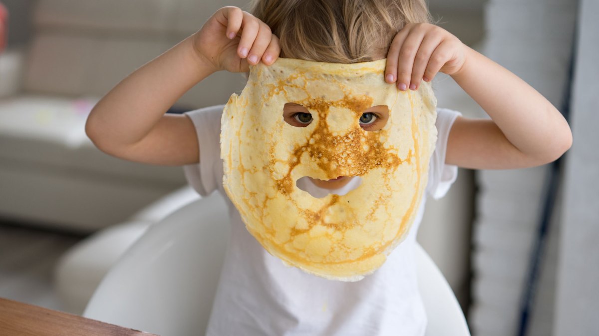Nahaufnahme eines kleinen Kindes, das am Küchentisch sitzt und sich einen Pfannkuchen vors Gesicht hält, in dem Löcher für Mund und Augen sind.