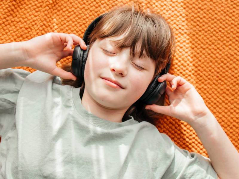 Mädchen liegt auf dem Fußboden, hat die Augen geschlossen und trägt On-ear-Kopfhörer.