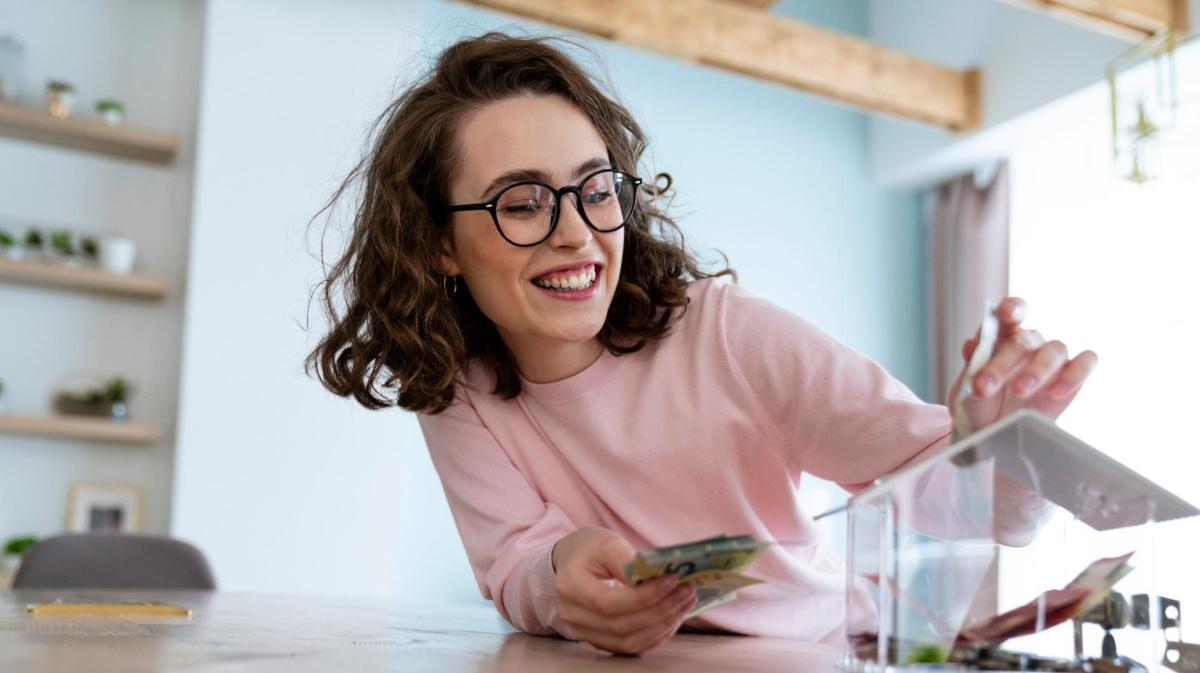 Frau mit Brille und rosa Shirt wirft Geld in eine Spardose und lacht.