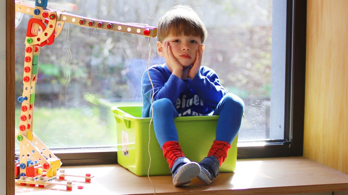 Kind sitzt in einer Spielkiste auf dem Fensterbrett und schaut gelangweilt.