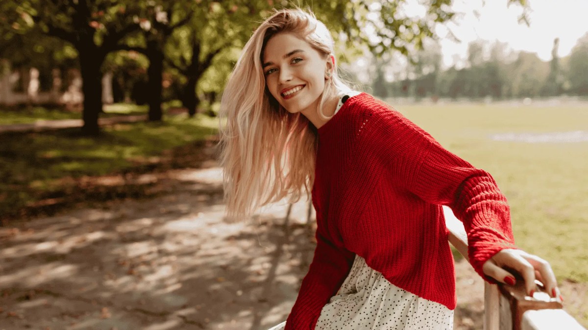Blonde Frau in rotem Pulli lächelt in Kamera, hinter ihr ein herbstlicher Park.