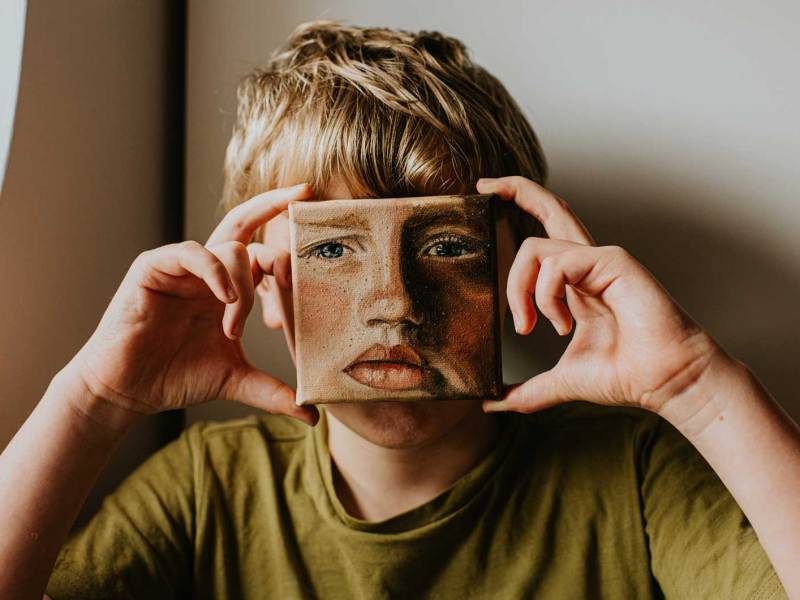 Junge hält sich ein kleines Ölgemälde von sich selbst vors Gesicht.