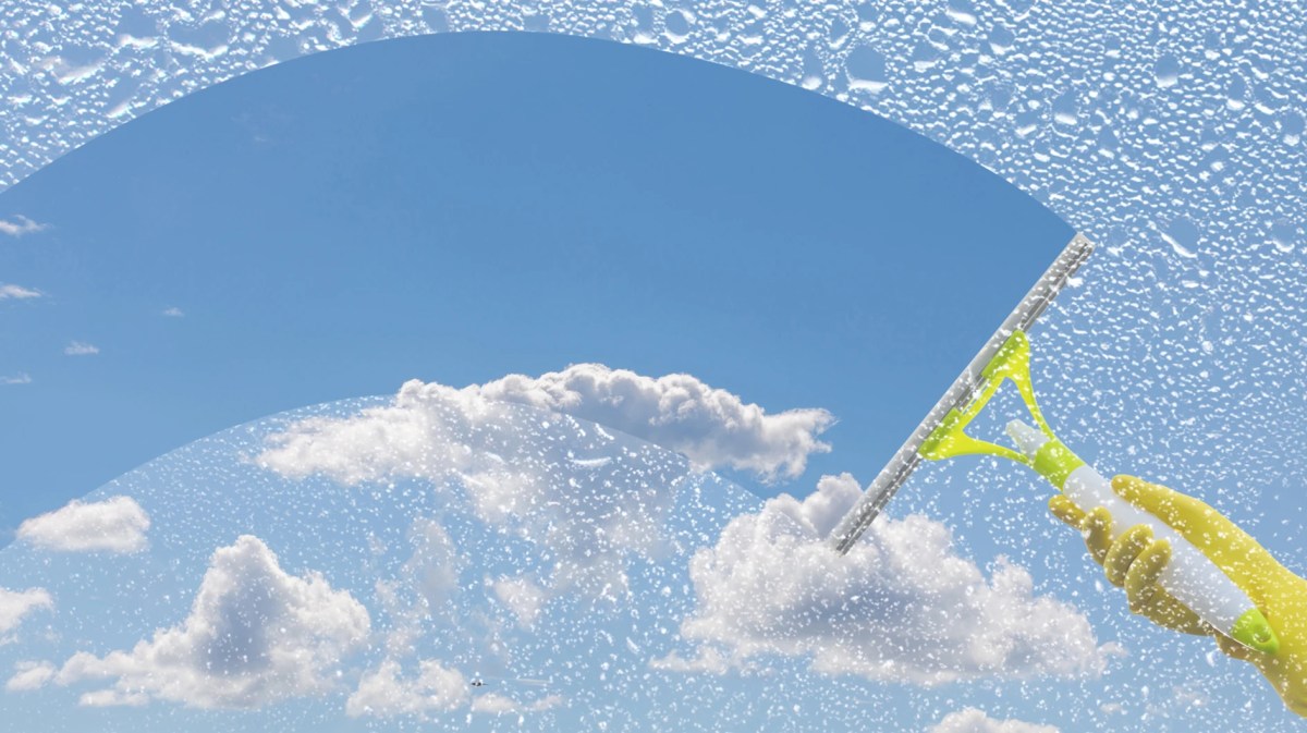 Hand in gelbem Gummihandschuh wischt mit Fensterabzieher Schaum von Scheibe und gibt den Blick auf blauen Himmel frei.