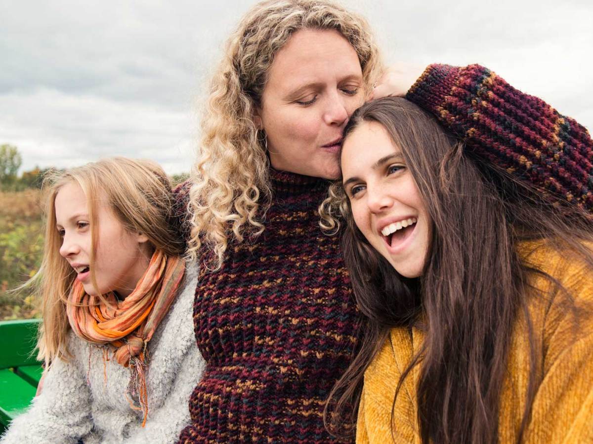 Mutter und zwei Töchter tragen warme Pullis und sitzen an einem bewölkten Herbsttag auf einer Bank in der Natur.