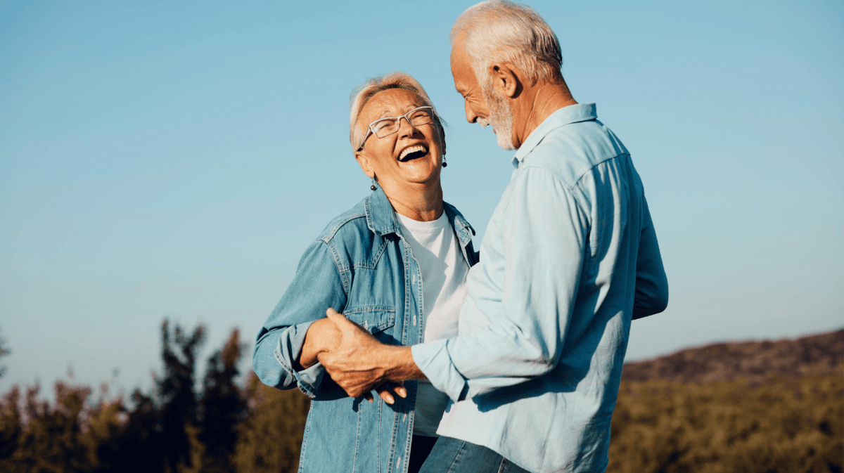 Älteres Paar steht in Feld, die Frau lacht beherzt, der Mann hält sie an den Händen.