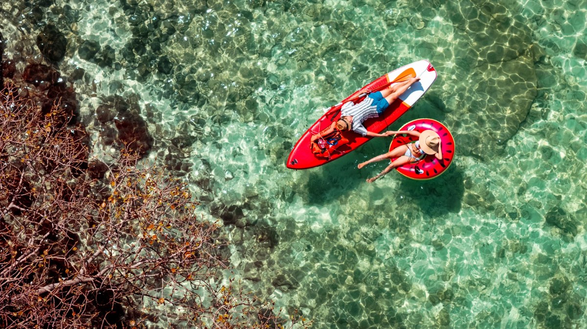 Ein junges Paar entspannt im klaren türkisgrünen Wasser, sie auf einer Wassermelonen-Luftmatratze, er auf einem roten SUP