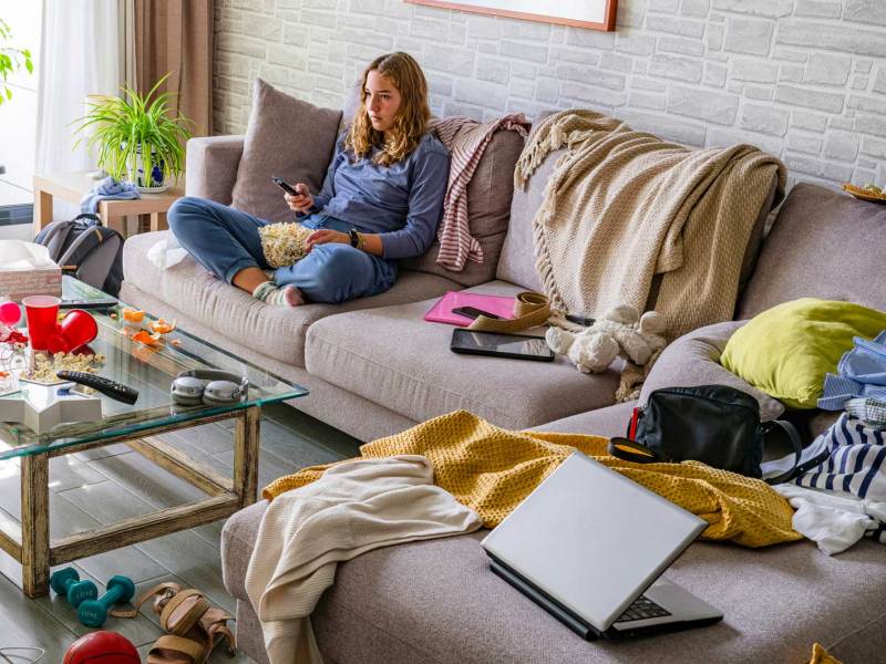 Mädchen im Teenageralter sitzt auf dem Sofa in einem unglaublich unordentlichen Wohnzimmer.