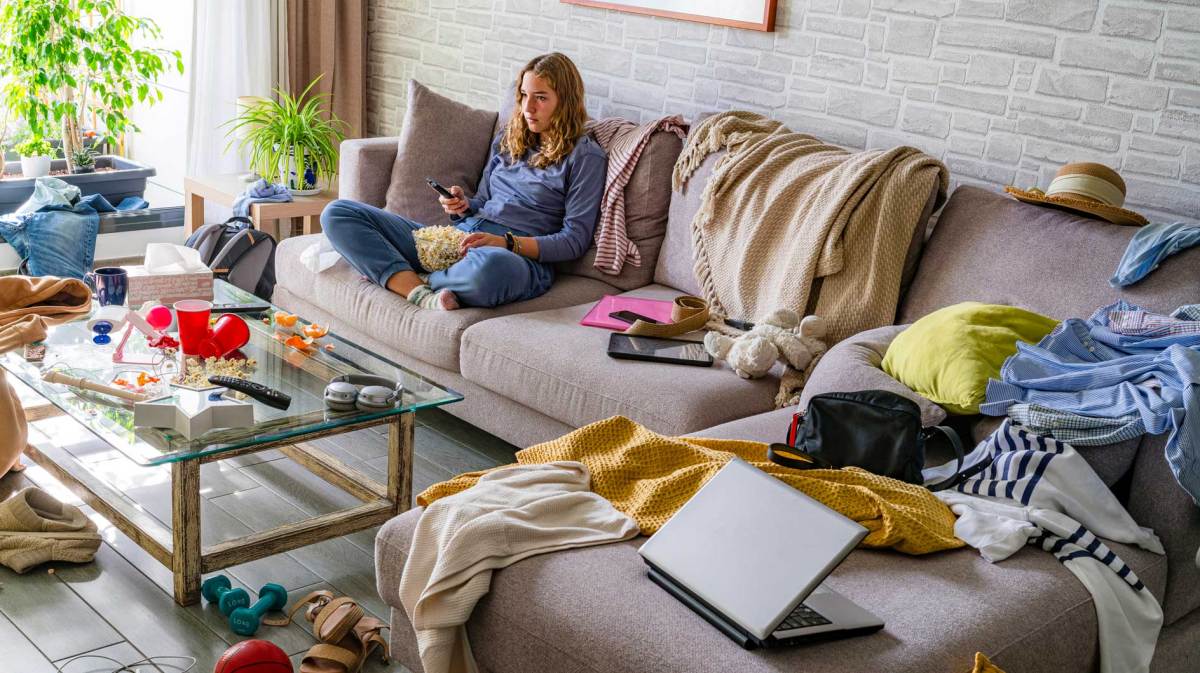 Mädchen im Teenageralter sitzt auf dem Sofa in einem unglaublich unordentlichen Wohnzimmer.