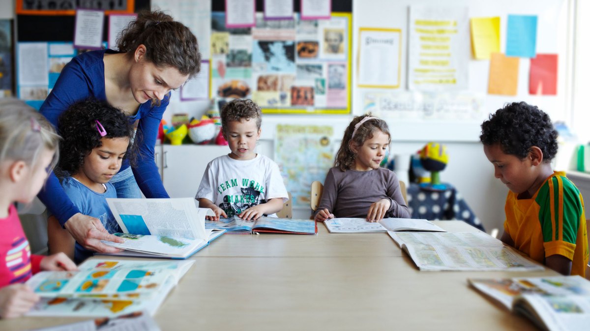 Gruppe Erstklässler sitzt mit Büchern an einem großen, runden Tisch, die Lehrerin beugt sich über eines der Kinder, um zu helfen.