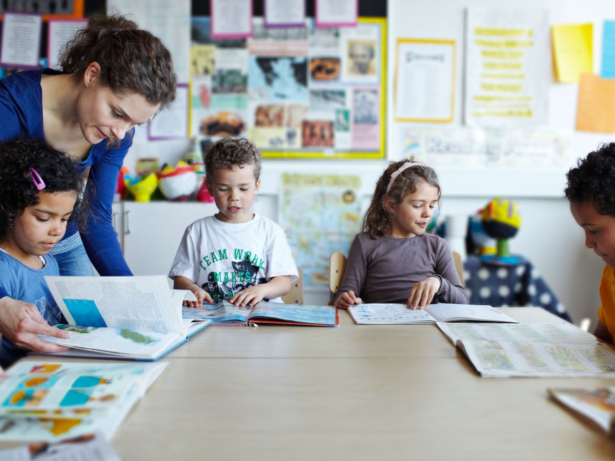 Gruppe Erstklässler sitzt mit Büchern an einem großen, runden Tisch, die Lehrerin beugt sich über eines der Kinder, um zu helfen.