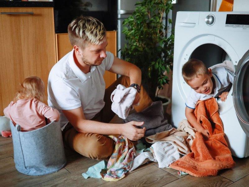 Vater sitzt vor der Waschmaschine auf dem Boden. Sein Sohn schaut aus der Wäschetrommel und die Tochter sitzt daneben in einem Stoffsack.