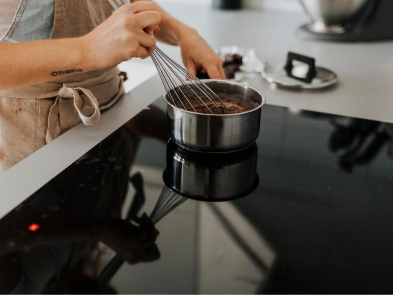 Eine Frau kocht eine Flüssigkeit im Topf auf einem Induktionsherd aus und rührt mit einem Schneebesen um.