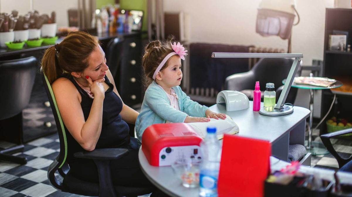 Kind sitzt auf dem Schoß der Mutter in einem Nagelstudio am Behandlungstisch.