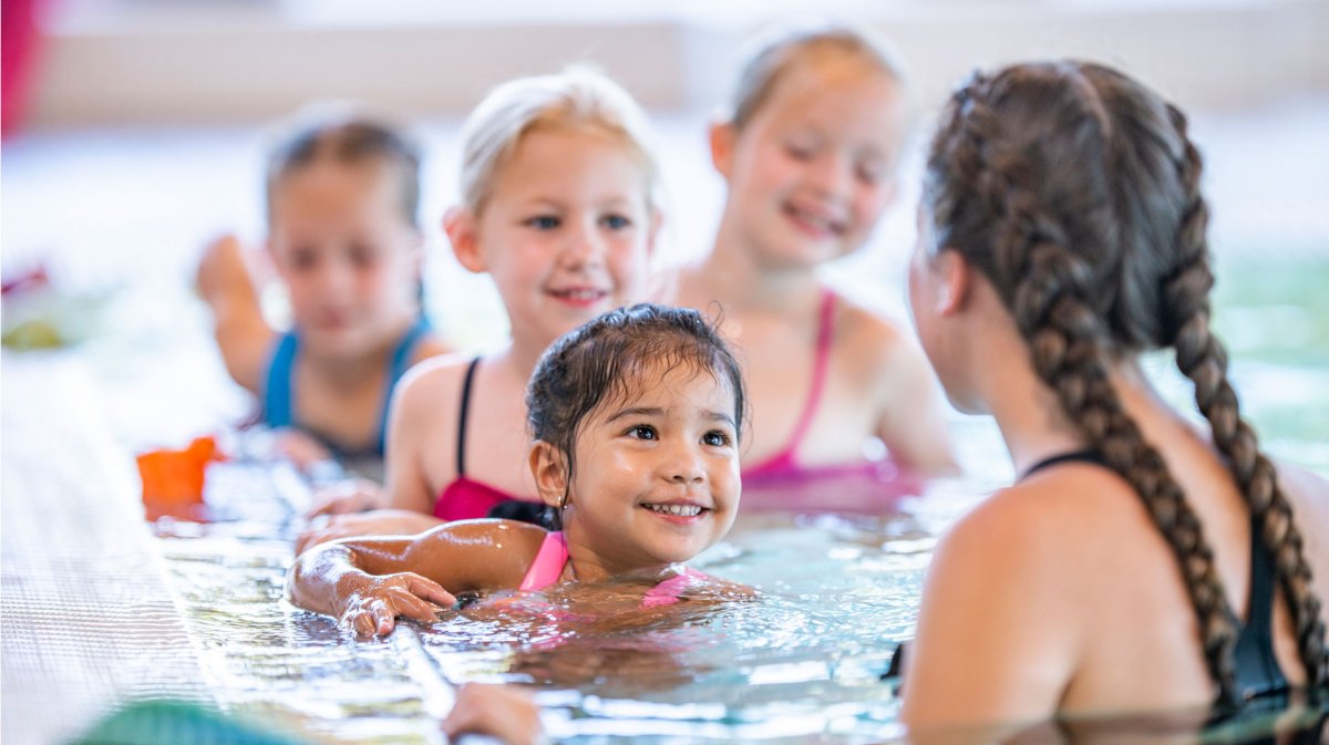 Gruppe von Schwimmschülerinnen im Becken. Die Schwimmtrainerin dreht der Kamera den Rücken zu.