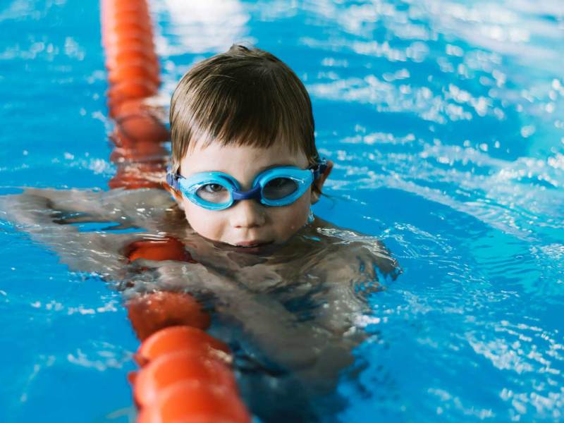 Junge, ca 6 Jahre alt, guckt mit Schwimmbrille aus dem Wasser eines Schwimmbads.