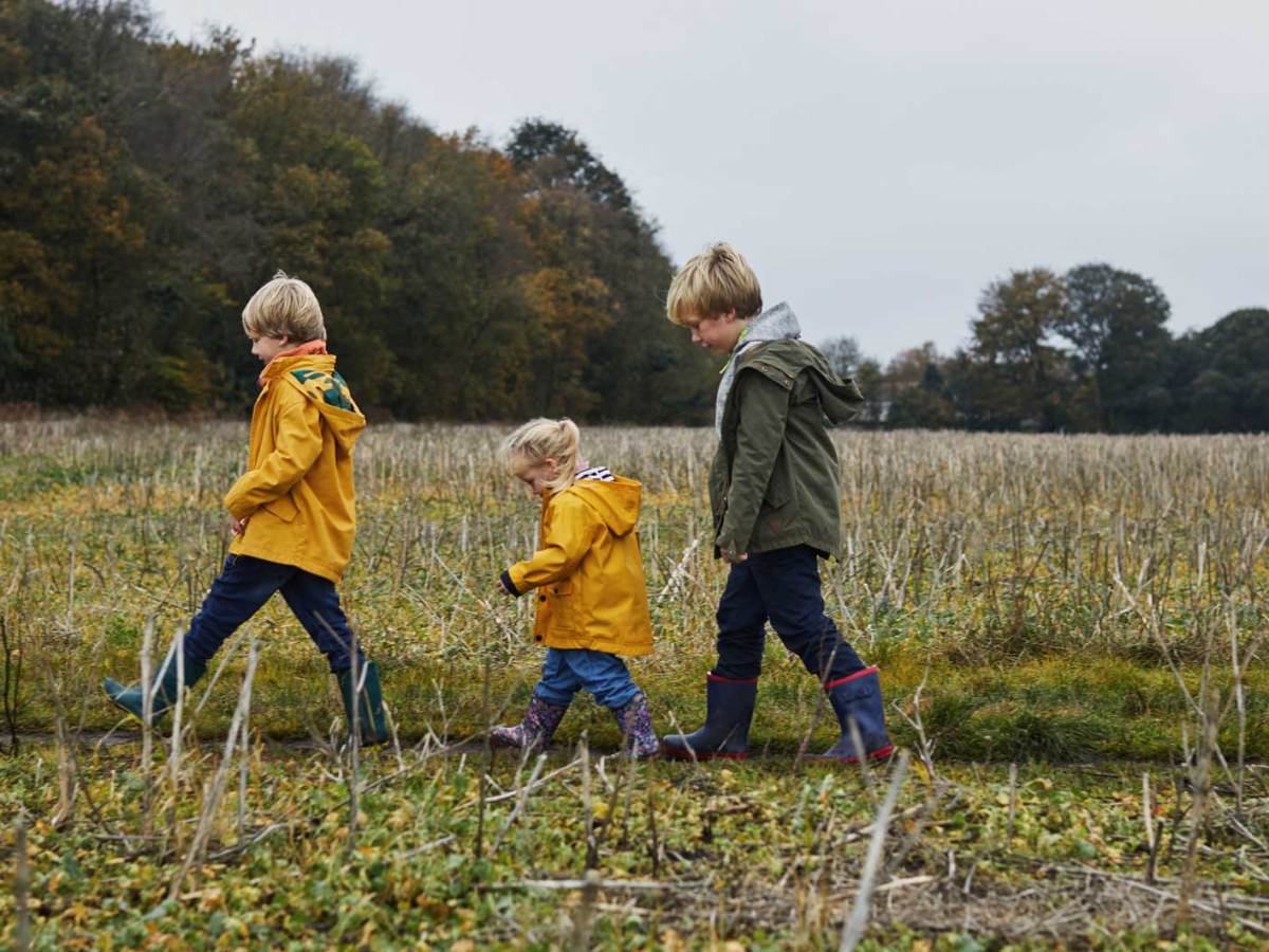 Drei Geschwister unterschiedlichen Alters gehen mit Gummistiefeln und Regenjacke einen Feldweg entlang.