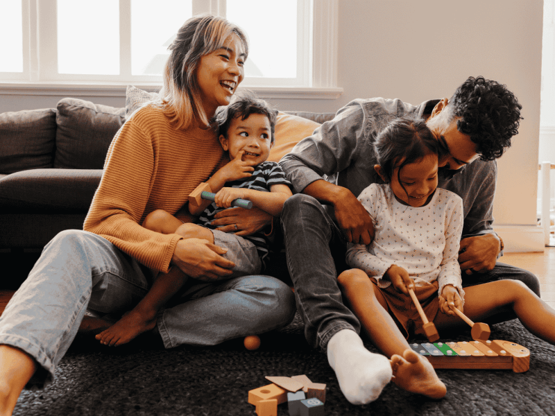 Familie mit zwei Kindern sitzt auf Boden vor Sofa und lacht.
