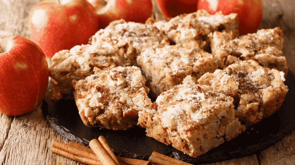 Apfel-Zimt-Kuchen auf einem Holztisch neben Äpfeln und Zimtstangen.