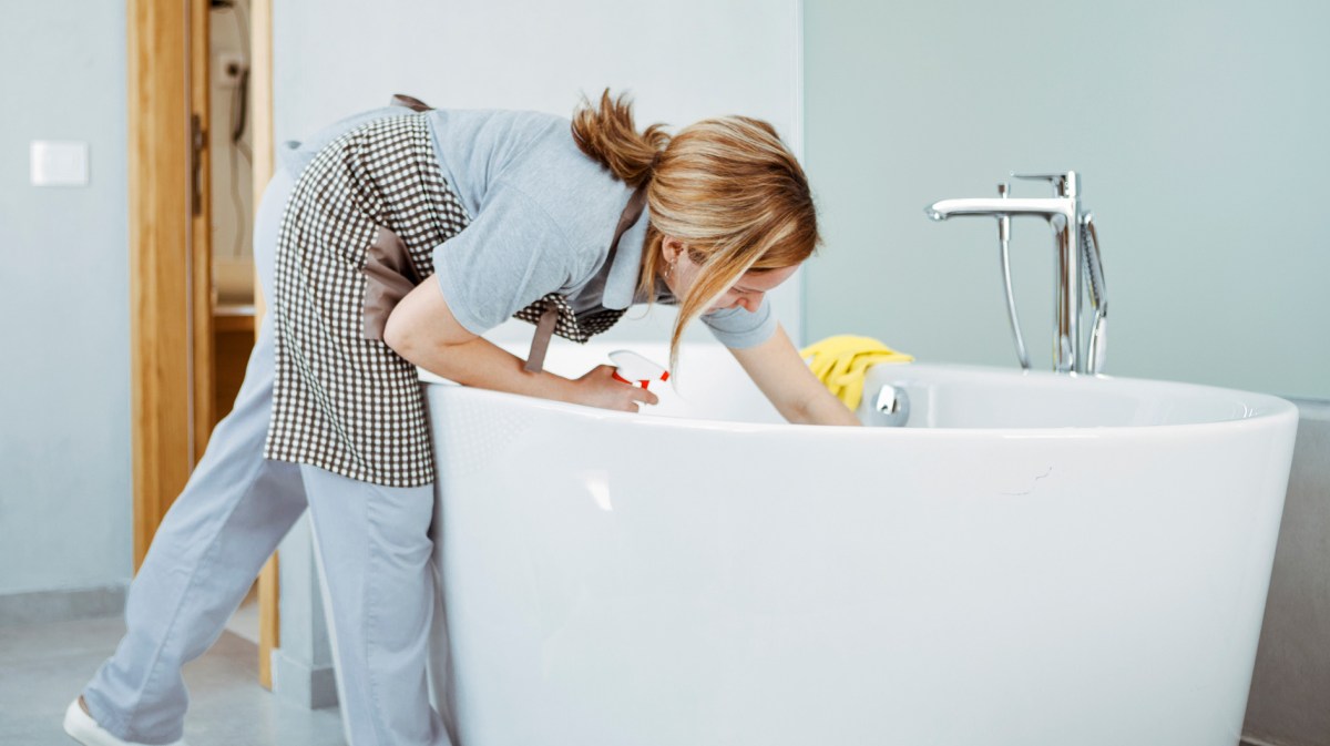 Frau putzt frei stehende Badewanne und trägt eine Schürze.