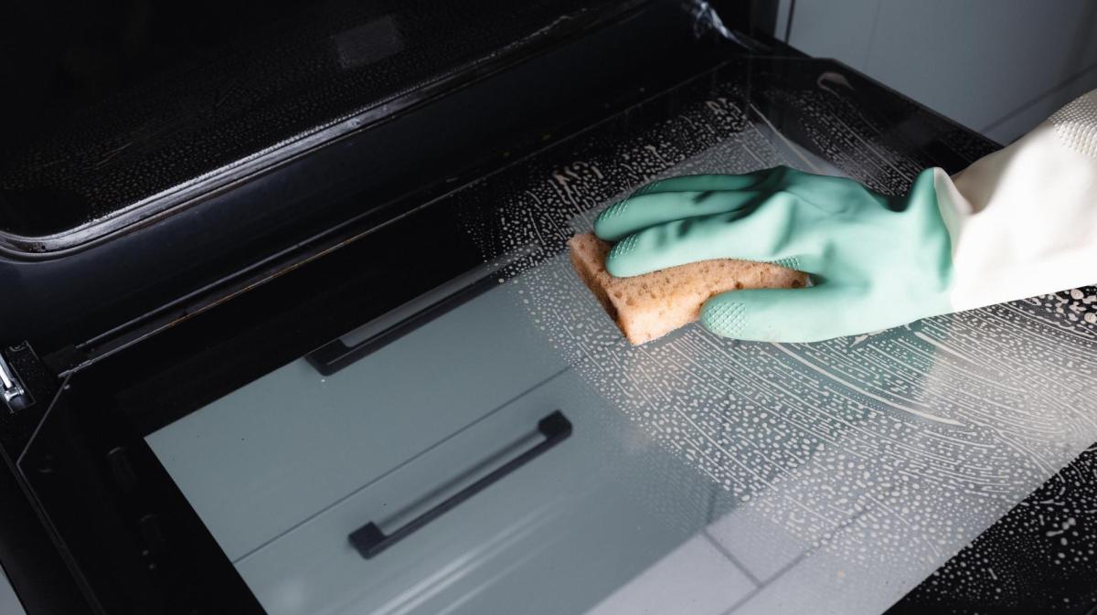 Person reinigt mit einem Handschuh und einem Schwamm den Backofen.