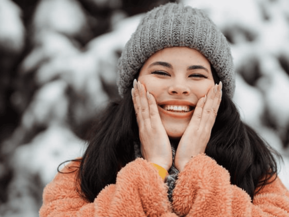 Junge Frau lächelt, trägt Mütze und Winterjacke, hinter ihr Schneebedeckte Bäume.