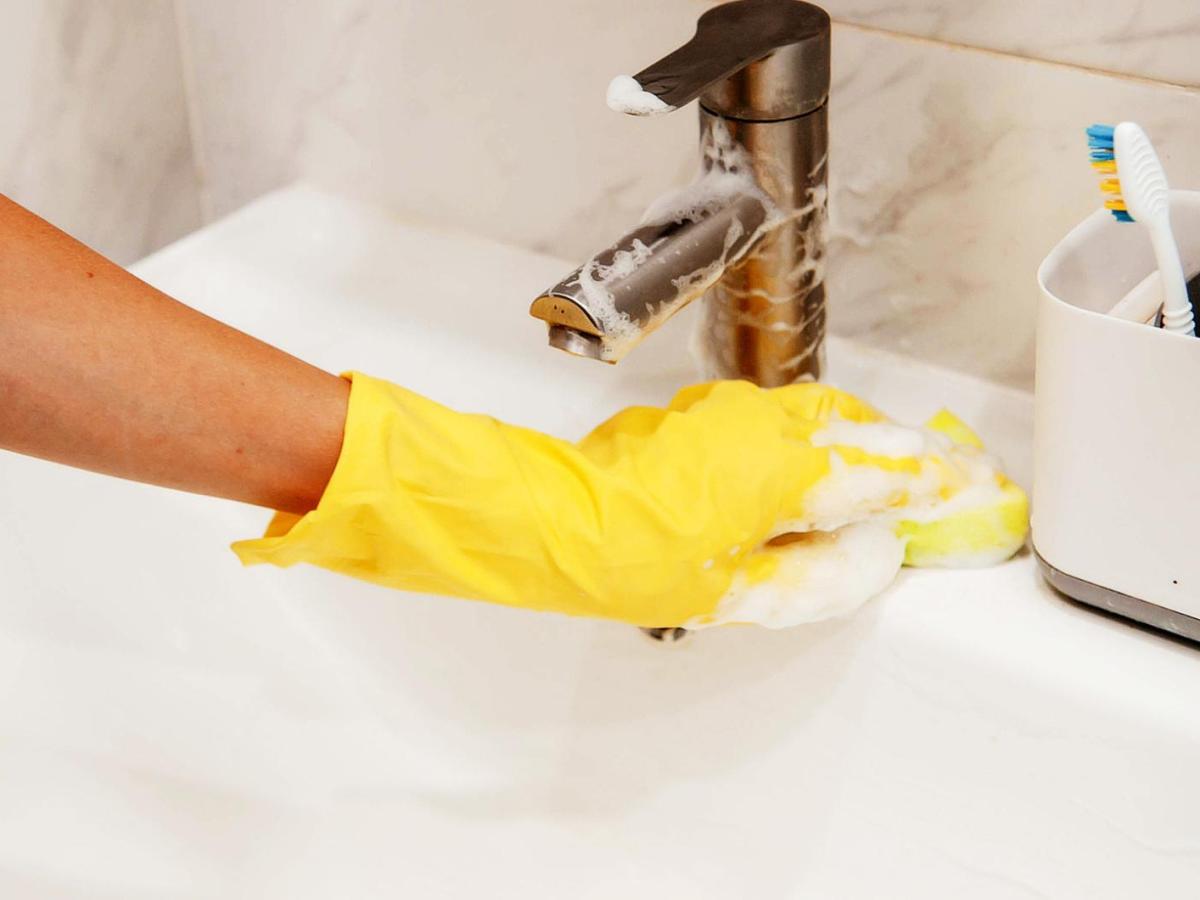 Frau mit gelben Handschuhen reinigt ein Waschbecken aus Keramik.