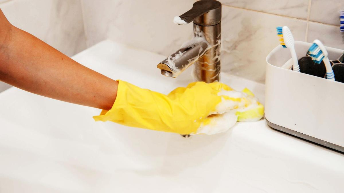 Frau mit gelben Handschuhen reinigt ein Waschbecken aus Keramik.