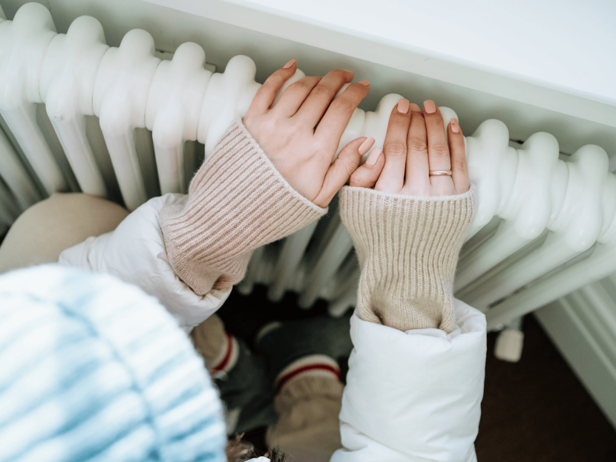 Frau sitzt warm angezogen vor der Heizung und hält ihre Hände auf die Heizung.