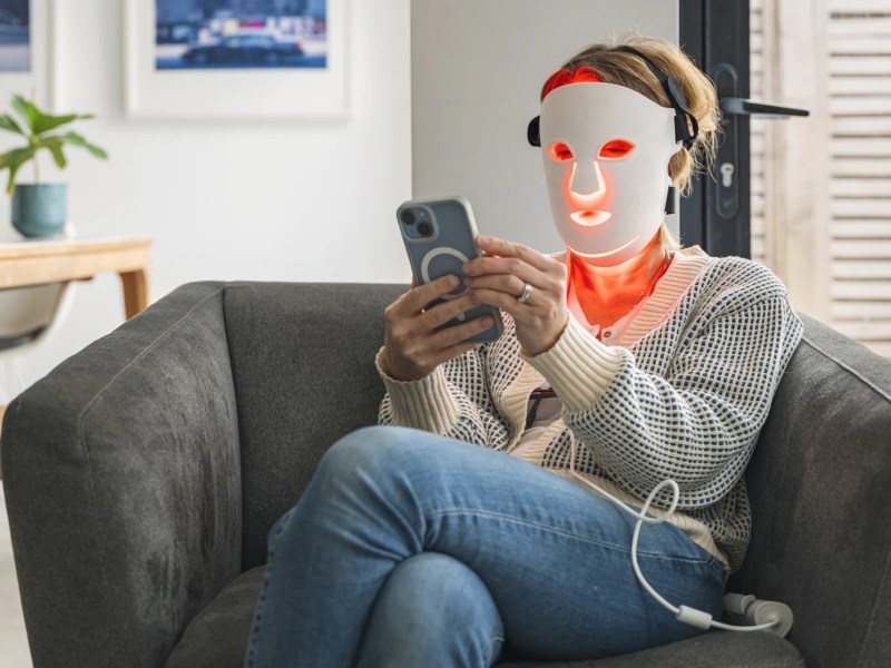 Frau nutzt LED-Gesichtsmaske zu Hause auf ihrem Sofa.