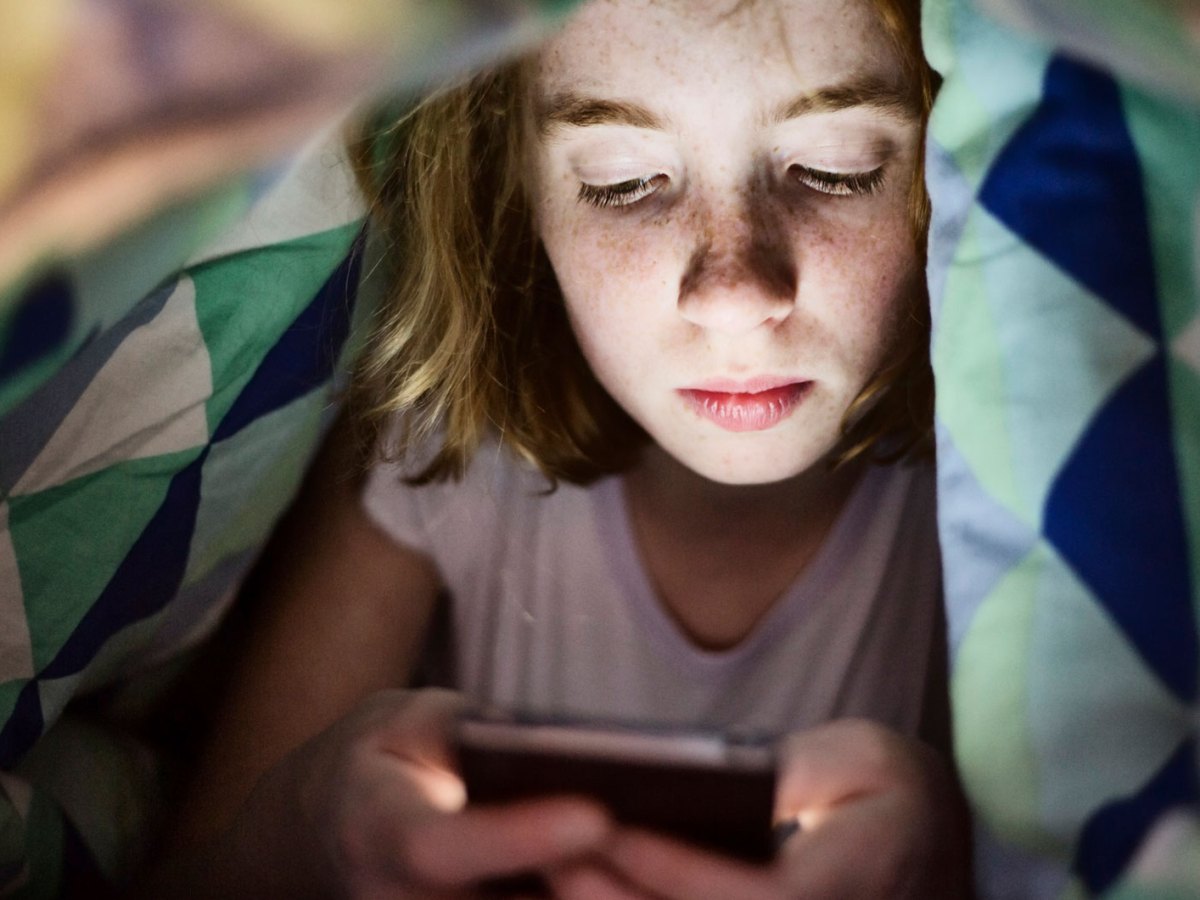 M&auml;dchen im Teenageralter liegt im Bett unter der Decke und schaut auf ihr Handy.