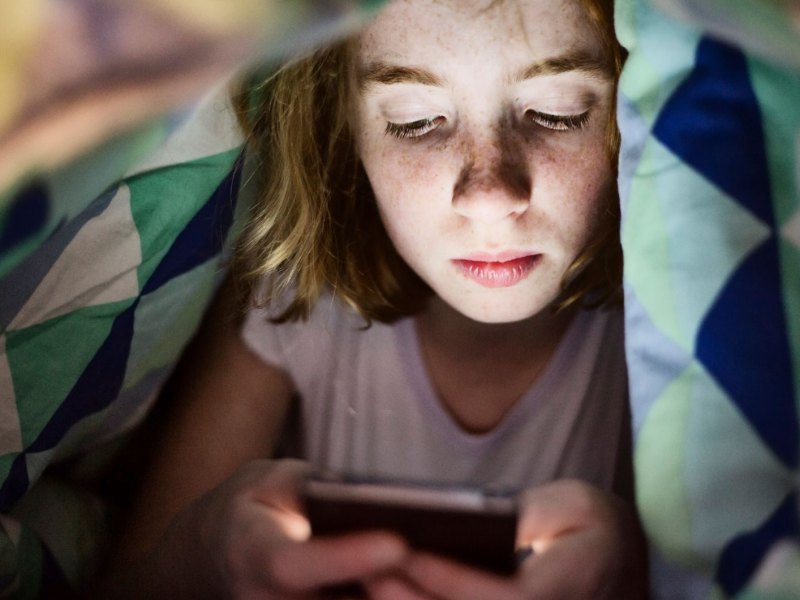 Mädchen im Teenageralter liegt im Bett unter der Decke und schaut auf ihr Handy.