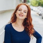 Hübsche Frau mit rotem Haar steht auf einem Gehweg und lächelt in die Kamera.