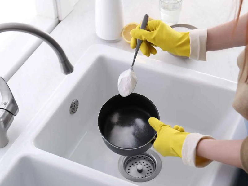 Frau mit Putzhandschuhen gibt Backpulver mit einem Esslöffel in eine Schüssel über dem Waschbecken.