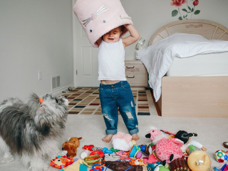 Kleinkind spielt im Zimmer und kippt einen Spielzeugkorb über seinem Kopf aus, sodass alles auf dem Boden liegt.
