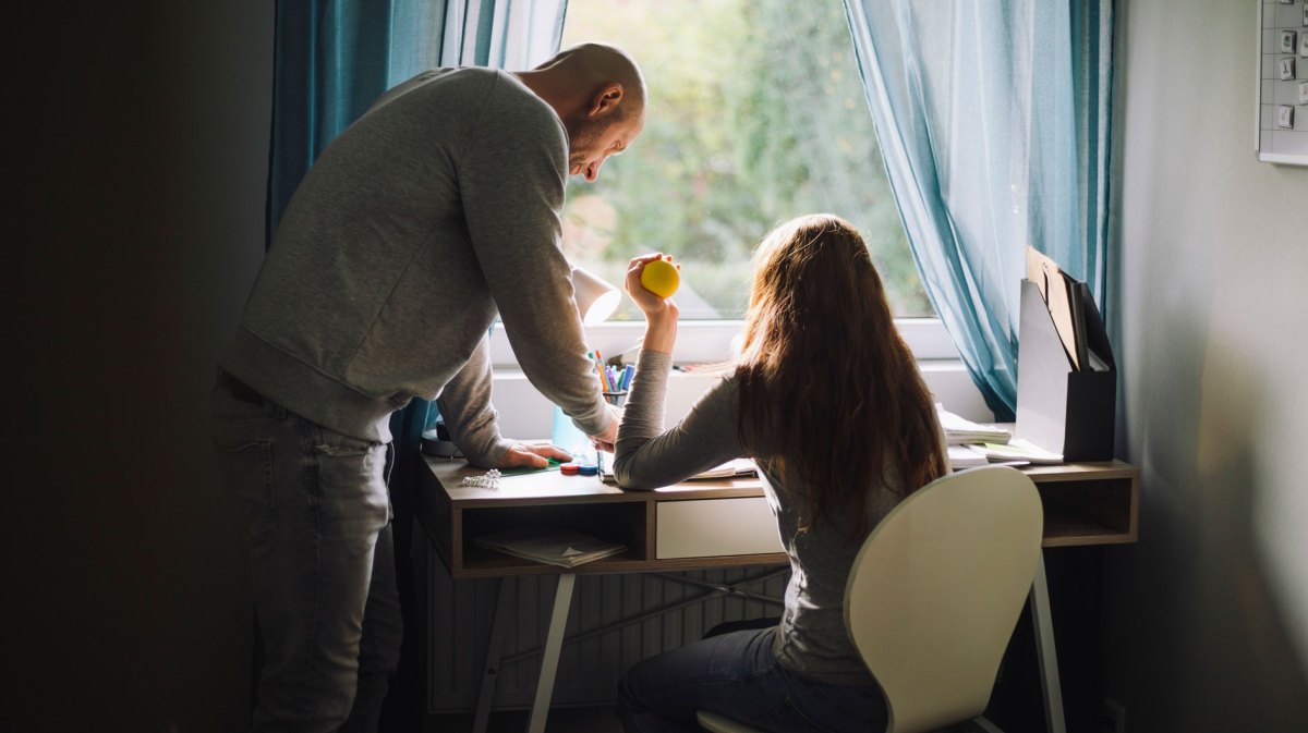Teenager-Mädchen sitzt mit Stressball in der Hand am Schreibtisch, sein Vater steht daneben und erklärt etwas.
