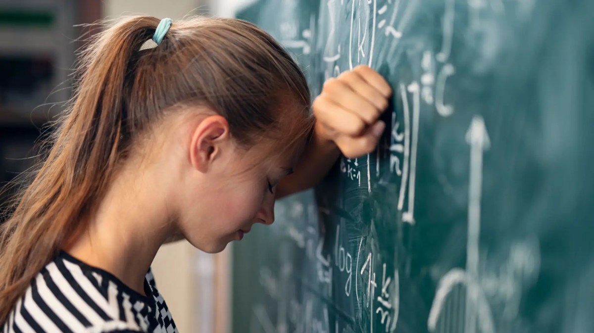 Mädchen lehnt mit Kopf an Tafel, auf der eine Matheaufgabe steht.